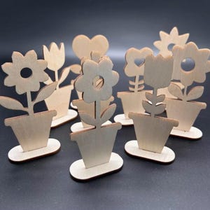 Könnte beinhalten: Eine Sammlung von Holz-Blumentopf-Ausschnitten. Jeder Ausschnitt zeigt ein anderes Blumendesign, darunter Gänseblümchen, Tulpen und eine herzförmige Blume. Die Töpfe stehen auf kleinen, ovalen Sockeln. Die natürliche Holzfarbe verleiht ihnen einen rustikalen Look.