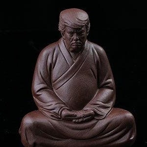 May include: A brown ceramic figurine of a seated figure in a meditative pose. The figure has a distinctive hairstyle and facial features. The figurine is set against a black background.