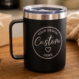 May include: A matte black insulated travel mug with a handle and a clear lid. The front features a circular white graphic with the text "YOUR DESIGN" at the top, "Custom" in script in the centre, a small heart icon below, and "HERE" at the bottom.