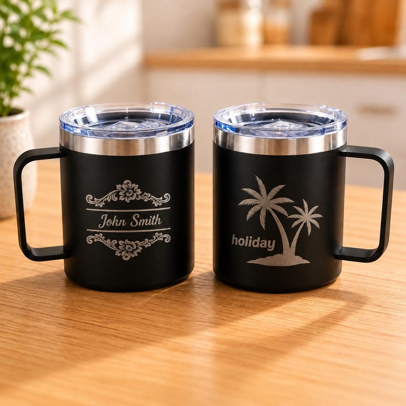 May include: Two black stainless steel mugs with clear lids and handles. The left mug features an engraved floral frame with the text John Smith. The right mug displays an engraved palm tree graphic above the word holiday in lowercase letters.