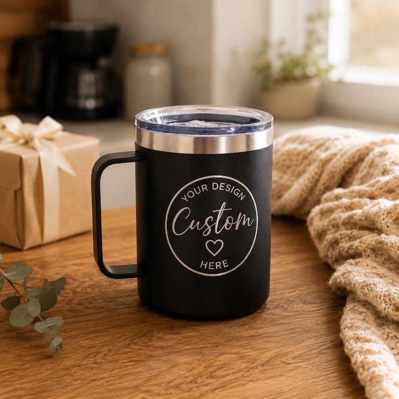 May include: A matte black insulated travel mug with a handle and a clear lid. The front features a circular white graphic with the text "YOUR DESIGN" at the top, "Custom" in script in the centre, a small heart icon below, and "HERE" at the bottom.