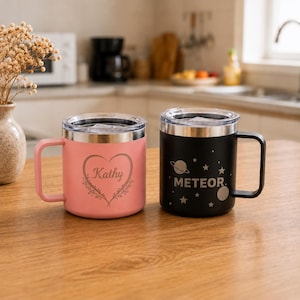 May include: Two stainless steel mugs with clear lids and handles. The pink mug features a heart design with the name Kathy engraved in the centre. The black mug features a space-themed design with stars, a planet, and the word METEOR engraved.
