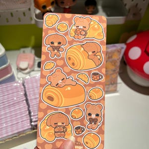 Op de afbeelding: Een stickervel met schattige cartoonberen en -puppy's, croissants en koffiekopjes. De stickers zijn in bruin- en oranje tinten, met de tekst "eries uage" onderaan. Het thema is een bakkerij of café.