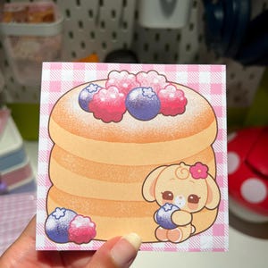 Peut inclure: Autocollant carré représentant une pile de crêpes garnies de myrtilles et de framboises. Un lapin de dessin animé avec une fleur tient une myrtille. Le sticker a une bordure rose à carreaux.