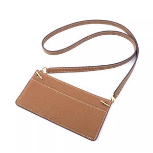 May include: A tan leather cardholder with a matching strap. The cardholder has a rectangular shape with white stitching and gold-colored hardware. The strap is thin and appears to be adjustable.