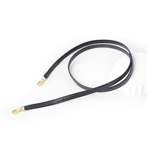 May include: Black leather bag strap with gold-colored metal clasps. The strap is thin and flexible, designed for use with handbags or other small bags. The strap is shown against a white background.