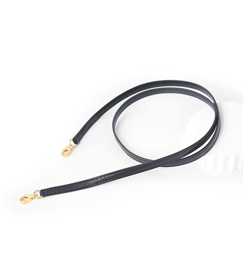 May include: Black leather bag strap with gold-colored metal clasps. The strap is thin and flexible, designed for use with handbags or other small bags. The strap is shown against a white background.