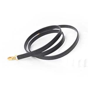 May include: A black leather purse strap with a gold-colored clasp. The strap is thin and flexible, designed to be attached to a handbag. The strap is shown against a white background.