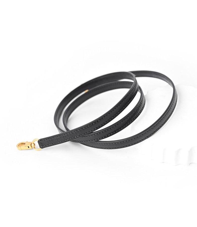 May include: A black leather purse strap with a gold-colored clasp. The strap is thin and flexible, designed to be attached to a handbag. The strap is shown against a white background.