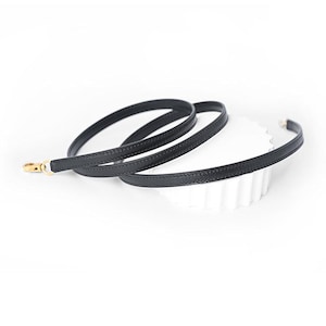 May include: A black leather strap with a gold-colored clasp. The strap is thin and appears to be stitched along the edges. The strap is coiled on a white surface.