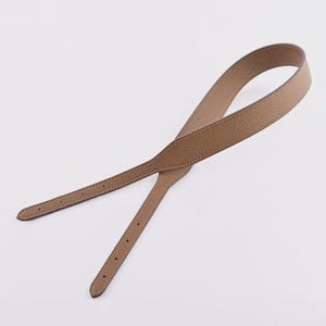 May include: A tan leather bag strap with a textured finish. The strap features a loop at one end and adjustment holes at the other. The strap is stitched with a lighter coloured thread.