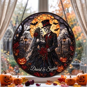 May include: A circular stained glass suncatcher featuring a skeleton couple in wedding attire, a large orange moon, bats, and pumpkins. The text David & Sophia is printed at the bottom. The piece is suspended by chains in a window with autumn foliage.