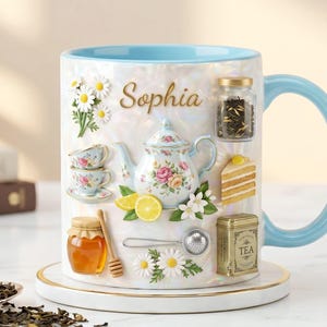 May include: A personalized ceramic mug with a light blue handle and rim, featuring a tea-themed design. The mug includes a teapot, teacups, lemons, honey, and the name "Sophia". Decorative elements include daisies and a slice of cake.