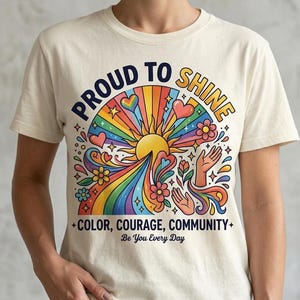 May include: A cream-coloured t-shirt featuring a colourful graphic with a central sun, rainbow rays, hearts, flowers, and two hands. The text reads: PROUD TO SHINE, COLOR, COURAGE, COMMUNITY, and Be You Every Day in a mix of bold and script fonts.