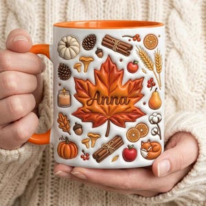 May include: A white ceramic mug with an orange interior and handle, featuring a large central maple leaf with the name Anna written in script. The design includes illustrations of pumpkins, cinnamon sticks, wheat, apples, and a fox.