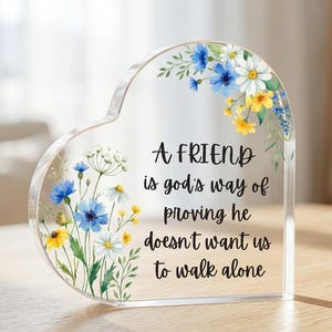 Puede incluir: Adorno decorativo en forma de corazón de acrílico transparente con detalles florales. El diseño presenta flores azules, amarillas y blancas y un mensaje impreso: "A FRIEND is god's way of proving he doesn't want us to walk alone."