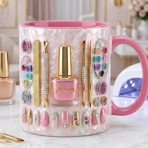 Könnte beinhalten: Eine rosa-weiße Tasse mit Nageldesign-Werkzeugen und einer Nagellackflasche mit dem Namen "Annie". Die Tasse ist mit Nagellack, Nagelkunst und Glitzer verziert. Der Griff ist rosa, und der Hintergrund deutet auf ein Salon-Ambiente hin.