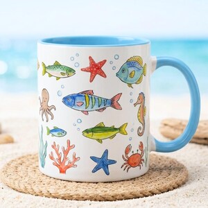 May include: White ceramic mug with a blue interior and handle, decorated with colorful illustrations of various sea creatures, including fish, starfish, and a seahorse. The mug is placed on a round, woven coaster, with a beach and ocean background.