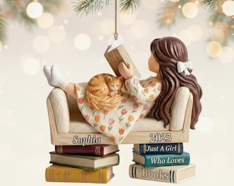 May include: A decorative ornament featuring a figure with long brown hair reading a book while reclining on a sofa with an orange cat. The sofa is inscribed with the name Sophia and the year 2025. The figure rests on a stack of books labeled Just A Girl Who Loves Books.