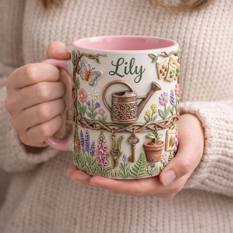 May include: A ceramic mug with a pink handle and interior, featuring a garden-themed design. The mug is adorned with a butterfly, flowers, a watering can, and the name "Lily". Decorative keys and a small potted plant are also visible.