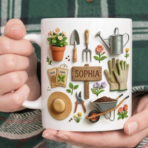 May include: White ceramic mug featuring a garden-themed design. The mug is decorated with gardening tools, flowers, seeds, a watering can, and a sign that reads "SOPHIA". The mug is held by a person.