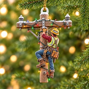 Op de afbeelding: Een kerstboomornament in de vorm van een houten elektriciteitspaal met een klimmende elektricien. De elektricien draagt een gele helm met de naam Logan, een rood geruit overhemd, een reflecterend veiligheidsvest, een spijkerbroek en bruine laarzen.