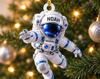 May include: A white and blue astronaut ornament featuring the name NOAH printed on the helmet visor. The astronaut is depicted in a floating pose with a reflective dark blue visor showing a space reflection, suspended by a gold string loop.