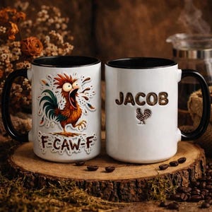 Pode incluir: Duas canecas de cerâmica brancas com alças e bordas pretas. Uma caneca apresenta um gráfico de galo de desenho animado com o texto "F-CAW-F". A outra caneca tem o nome "JACOB" e um gráfico de galo. As canecas estão em uma superfície de madeira com grãos de café.