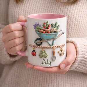 May include: White ceramic mug with a pink interior and handle, featuring a garden-themed design. A blue wheelbarrow holds plants and tomatoes. Charms of a ladybug, gloves, shovel, and bee hang below. The name "Lily" is written on the mug.