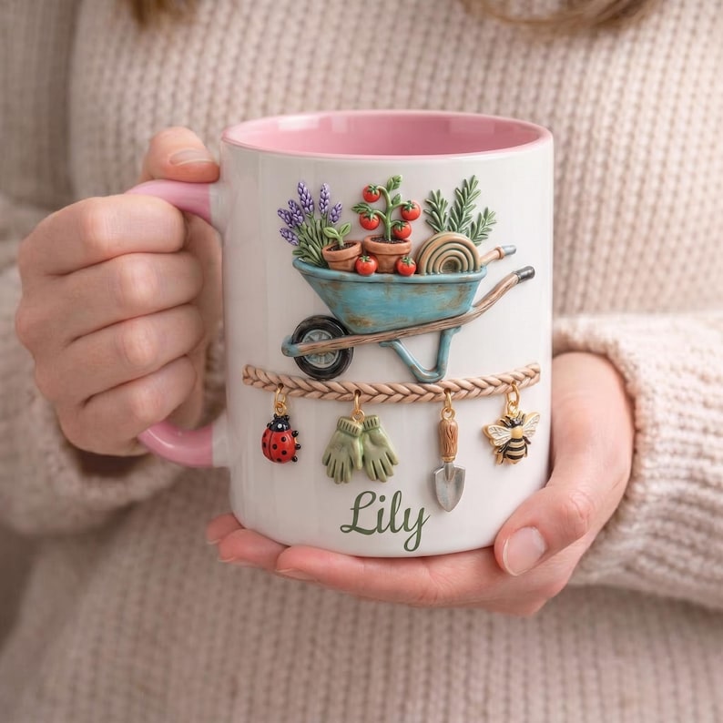 May include: White ceramic mug with a pink interior and handle, featuring a garden-themed design. A blue wheelbarrow holds plants and tomatoes. Charms of a ladybug, gloves, shovel, and bee hang below. The name "Lily" is written on the mug.