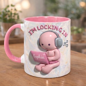 Puede incluir: Taza de cerámica blanca con interior y asa rosas. La taza presenta una figura rosa con auriculares y sosteniendo un portátil con el nombre "Sophia". El texto "I'M LOCKING IN" está impreso en la taza, junto con notas musicales.