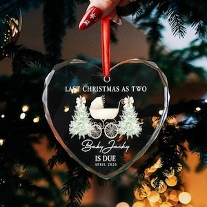 Op de afbeelding: Hartvormige glazen kerstversiering met de tekst "Last Christmas as Two" en "Baby Jacky is Due April 2026". De versiering heeft een kinderwagen en twee kerstbomen. Een rood lint is bevestigd om op te hangen.