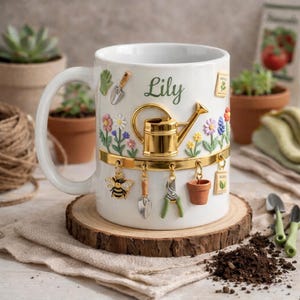 May include: White ceramic mug with a garden theme. The mug features a gold watering can, a bee charm, and miniature gardening tools. The name "Lily" is written in green script. The mug is decorated with colorful flowers and sits on a wooden slice.