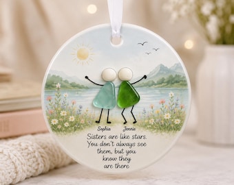 May include: A round white ceramic ornament hanging from a white ribbon. It features a printed landscape with a lake, mountains, and flowers. Two figures made of sea glass in light blue and green with painted black limbs stand in the foreground.