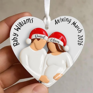 Op de afbeelding: Hartvormige kerstversiering met een stel met kerstmutsen. De versiering leest "Baby Williams Arriving March 2026" met de namen "Daniel" en "Olivia" op de mutsen. De versiering is wit met een wit lint.