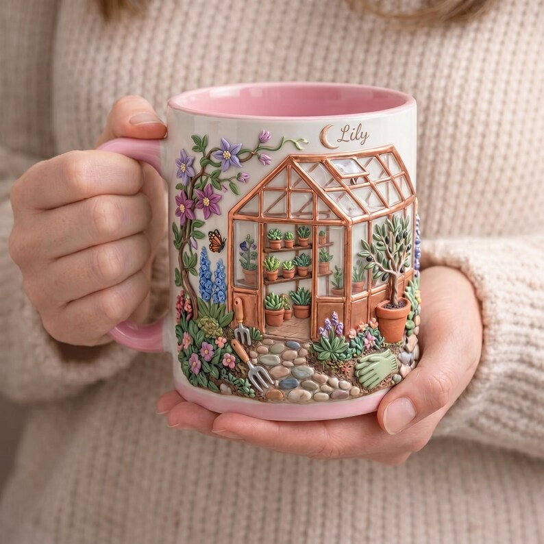 May include: A white ceramic mug with a pink interior and handle, featuring a detailed 3D greenhouse design. The greenhouse is filled with potted plants and surrounded by colorful flowers, a butterfly, and gardening tools. The name "Lily" is written on the mug.