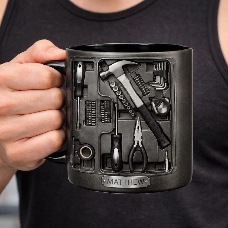 K&ouml;nnte beinhalten: Eine schwarze Keramik-Tasse mit Werkzeugkasten-Design, das gepr&auml;gte Werkzeuge wie Hammer, Schraubendreher und Zangen zeigt. Der Name "MATTHEW" ist auf einer Plakette zu sehen. Die Tasse wird von einer Person gehalten.