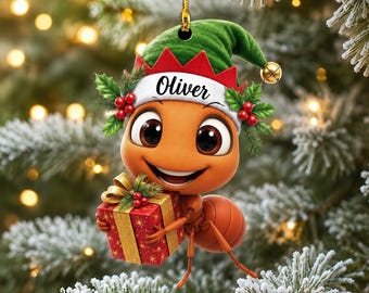 May include: A hanging Christmas ornament featuring a cartoon ant wearing a green and red elf hat with the name Oliver printed on the white brim. The ant has large eyes, a wide smile, and is holding a red gift box with a gold ribbon and holly.