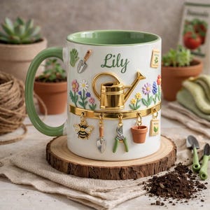 May include: White ceramic mug with a green handle and rim, decorated with gardening-themed charms and the name "Lily". Features a gold watering can, flowers, a bee, and miniature gardening tools. The mug sits on a wooden slice.