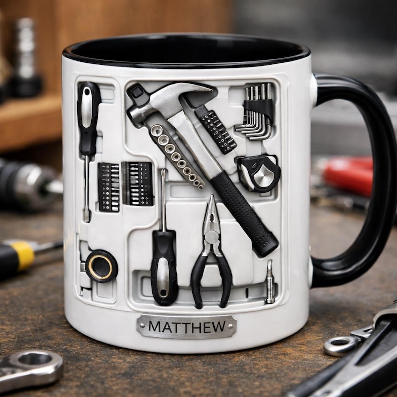 K&ouml;nnte beinhalten: Wei&szlig;er Keramikbecher mit schwarzem Innenraum und Henkel, mit einem Werkzeugkasten-Design. Der Becher zeigt einen Hammer, Schraubendreher, Zangen und andere Werkzeuge. Eine kleine Plakette unten zeigt "MATTHEW".