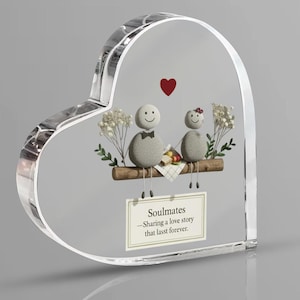 May include: Heart-shaped clear acrylic keepsake with a decorative design. The design features two pebble figures with smiling faces, a red heart, and the text "Soulmates - Sharing a love story that lasts forever."