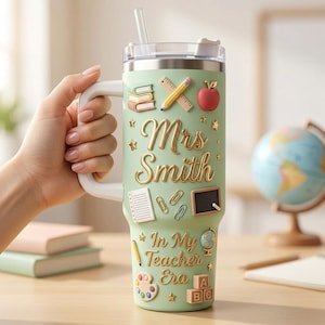May include: A light green insulated tumbler with a handle and clear lid featuring raised school-themed icons including books, a pencil, an apple, a chalkboard, and a paint palette. The text reads Mrs Smith In My Teacher Era in gold script.