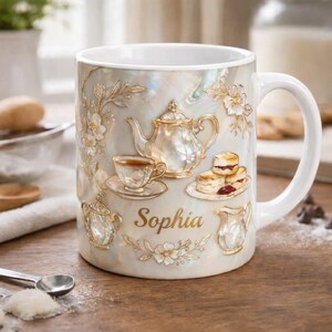 May include: White ceramic mug with a pearlescent finish, featuring embossed gold-colored tea party details. The design includes a teapot, teacup, scones, and the name "Sophia". The mug has a white handle.