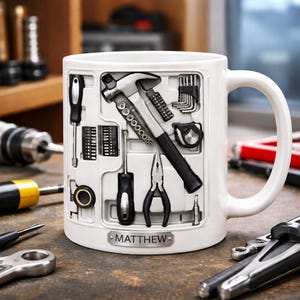 K&ouml;nnte beinhalten: Wei&szlig;e Keramiktasse mit Werkzeugkasten-Design. Die Tasse zeigt einen Hammer, Schraubendreher, Zangen und andere Werkzeuge. Ein kleines Metallschild mit dem Namen "MATTHEW" befindet sich auf der Tasse. Die Tasse steht auf einer Holzoberfl&auml;che.