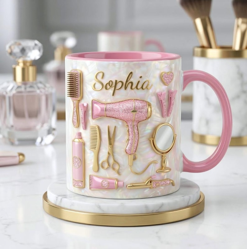 May include: A white mug with a pink handle and interior, featuring the name "Sophia" in gold script. The mug is decorated with pink and gold hair styling tools, including a hairdryer, comb, and scissors. It sits on a marble coaster with a gold rim.