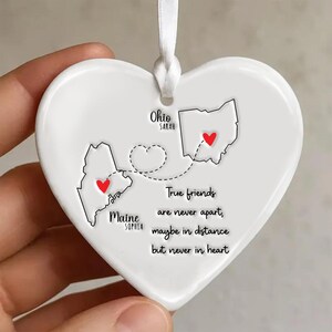 May include: White heart-shaped ornament with a ribbon for hanging. It features outlines of the states of Ohio and Maine, each with a red heart. Text reads "True friends are never apart, maybe in distance but never in heart."