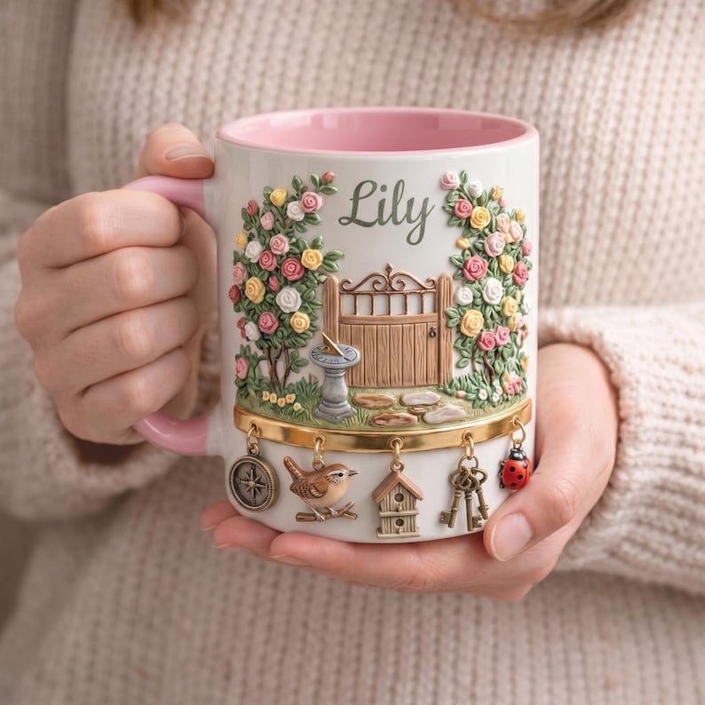 May include: A white ceramic mug with a pink interior and handle, featuring a garden scene with roses, a gate, and the name "Lily." Decorative charms include a compass, bird, birdhouse, keys, and a ladybug.