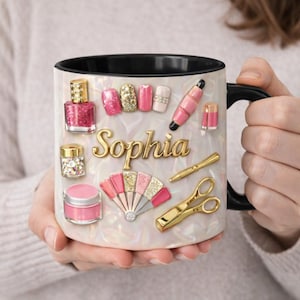 Op de afbeelding: Een witte koffiemok met een zwarte binnenkant en handvat, versierd met nail art en manicure gereedschap. De mok toont de naam "Sophia" in gouden letters, samen met nagellakflesjes, glitter en een waaierontwerp.