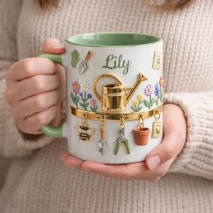 May include: White ceramic mug with a green handle and rim, featuring a garden theme. The mug is decorated with a gold watering can, flowers, and hanging charms of gardening tools and a bee. The name "Lily" is written on the mug.