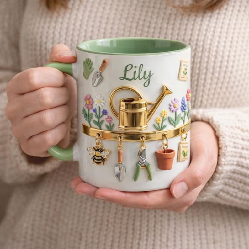 May include: White ceramic mug with a green handle and rim, featuring a garden theme. The mug is decorated with a gold watering can, flowers, and hanging charms of gardening tools and a bee. The name "Lily" is written on the mug.